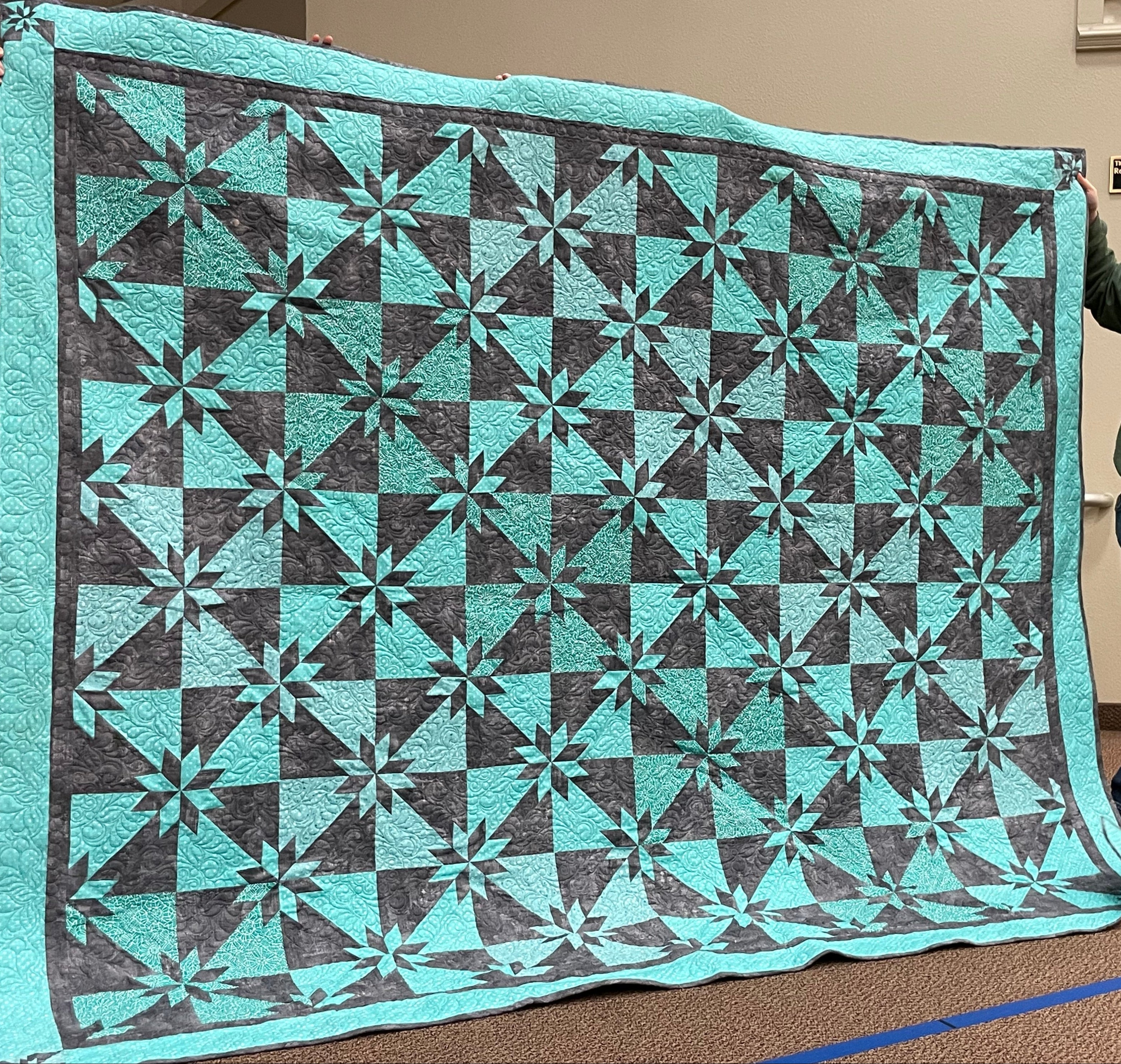 Person holding a large teal and gray quilt with a hunters star pattern in an indoor setting.